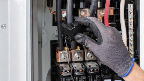 Gloved hand clamping a split-core current transformer around a conductor inside an open electrical panel