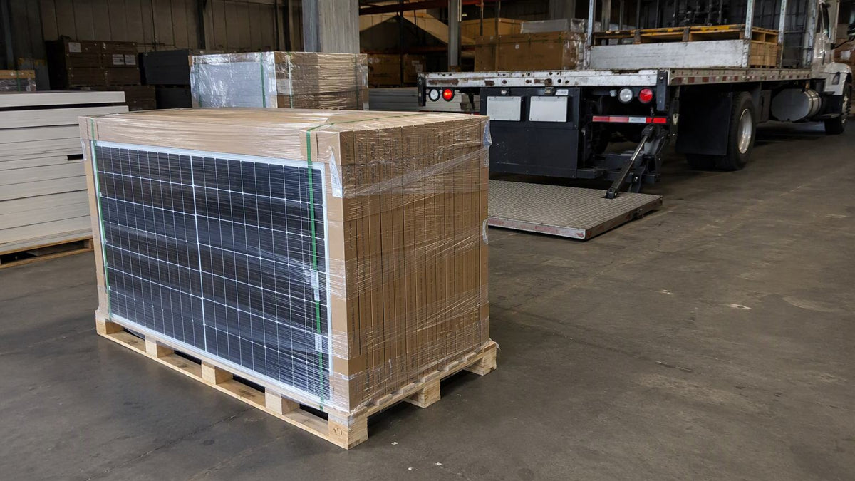 Full wooden pallet stacked with boxed solar panels in shrink film on a warehouse floor with a flatbed liftgate truck behind