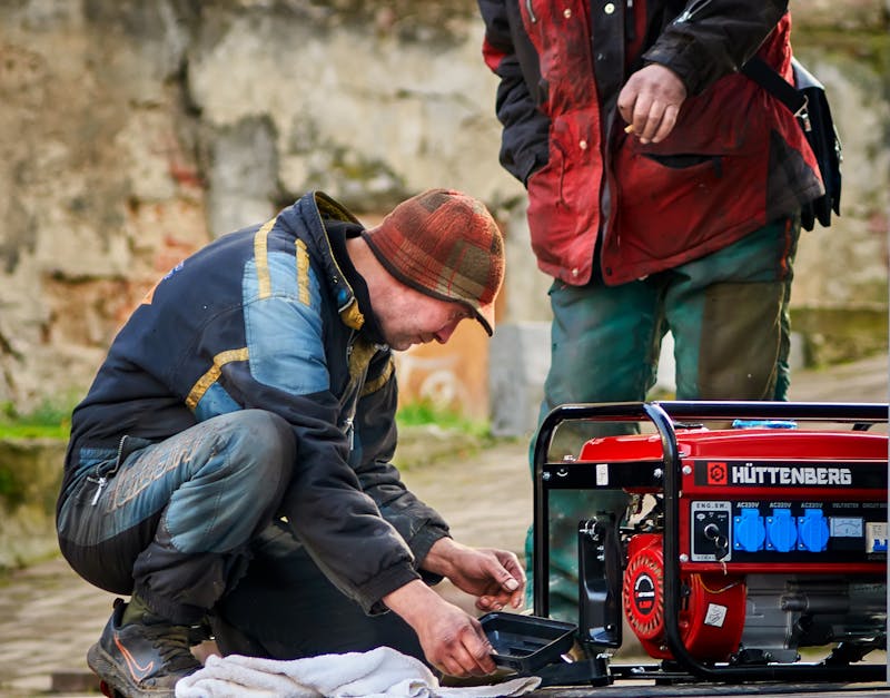 Two men repairing a red portable generator outdoors on a stone pavement