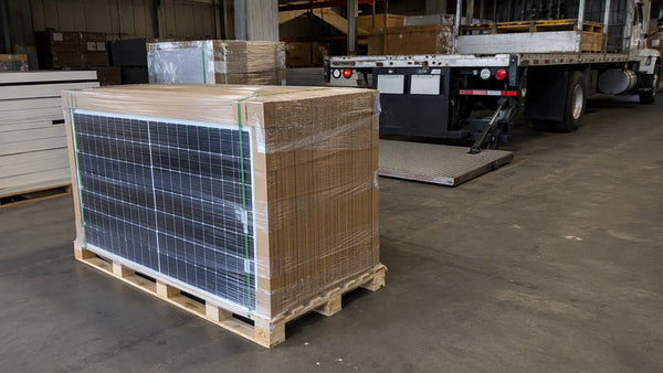 Full wooden pallet stacked with boxed solar panels in shrink film on a warehouse floor with a flatbed liftgate truck behind